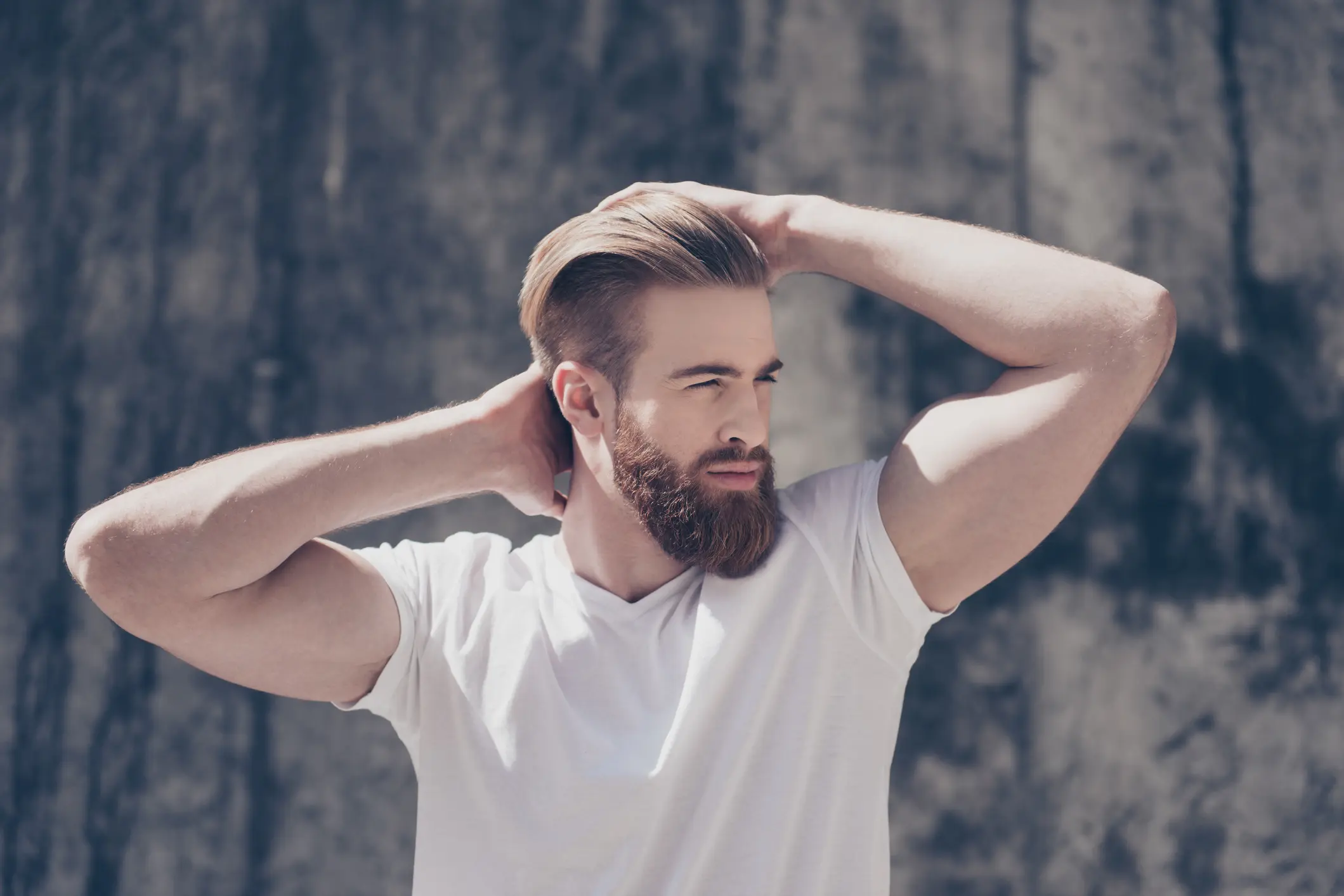 Bearded man in white t-shirt posing outdoors with hands behind head.